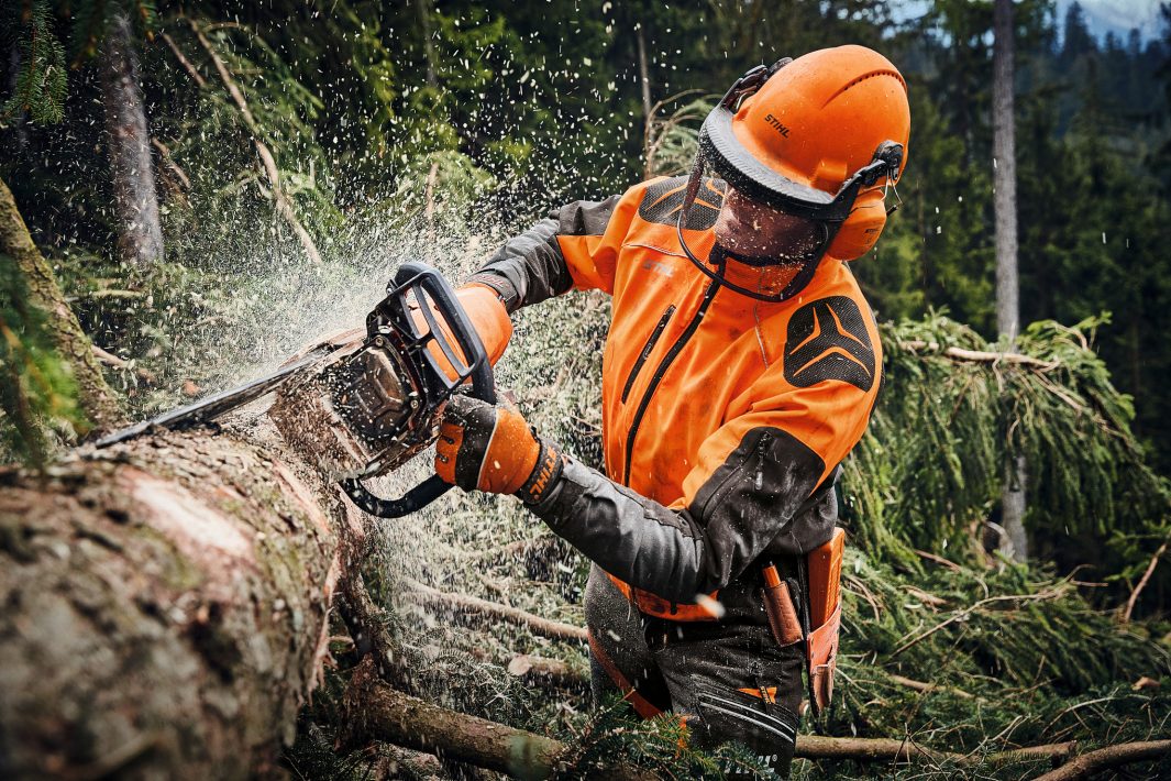 Person in Stihl orange safety gear cutting a tree with a chainsaw in a forest setting
