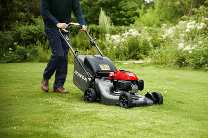 A Honda walk-behind lawn mower being pushed across a green grass lawn by a person whose legs and hands are visible. The mower has a black body with a red engine cover and black wheels.