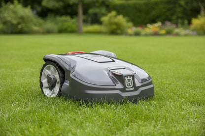 A Husqvarna robotic lawnmower in dark gray and black with a red emergency stop button on top, shown from a three-quarters angle - From Morgans Machinery