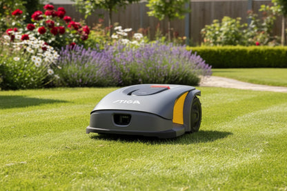 A STIGA  robotic lawnmower in gray and yellow is shown on a green grass lawn in a sunny garden with flowers and a fence in the background.