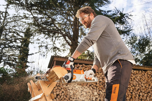 Man using a stihl chainsaw to cut wood with trees and stacked logs in the background