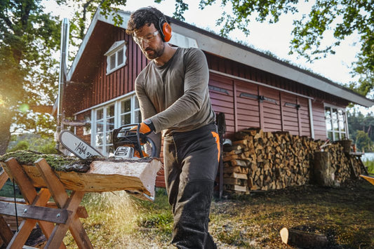 Man using a stihl chainsaw to cut wood in front of a cabin.