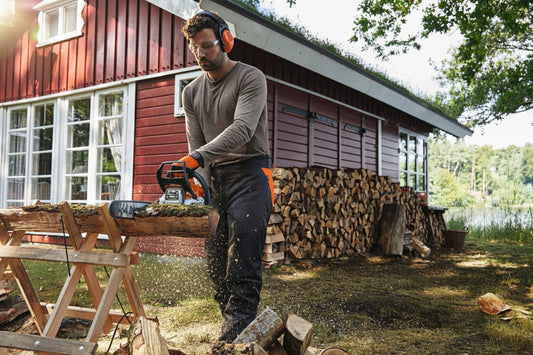 Man using a stihl chainsaw to cut wood in front of a red cabin.