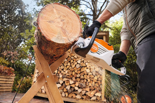 Person using a stihl chainsaw to cut wood with a log and stacked firewood in the background.